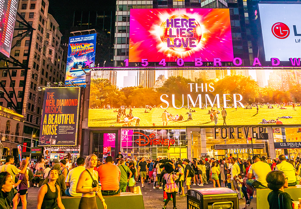 Two digital signs in Times Square above the Disney Store. The upper sign is the focus of the image and has an advertisement for Here Lies Love.