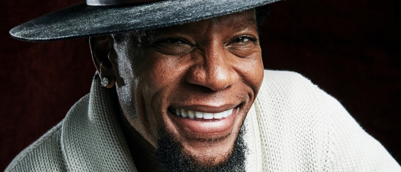 D.L. Hughley wearing a hat and grinning at the camera