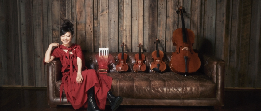 Hiromi wearing a red dress and sitting on a sofa next to four stringed instruments of varying sizes