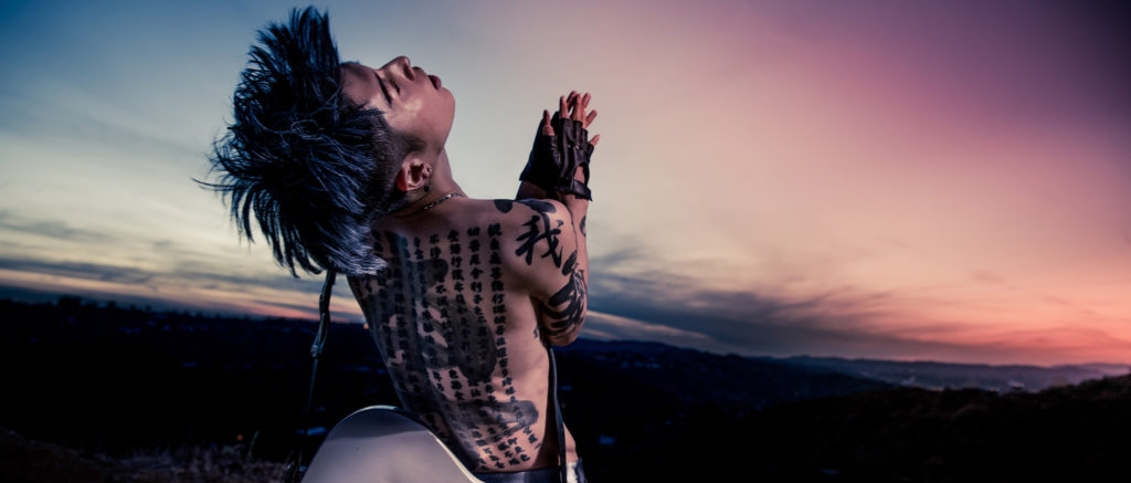 Miyavi stands shirtless with a guitar slung over his back in front of a sunset on the horizon