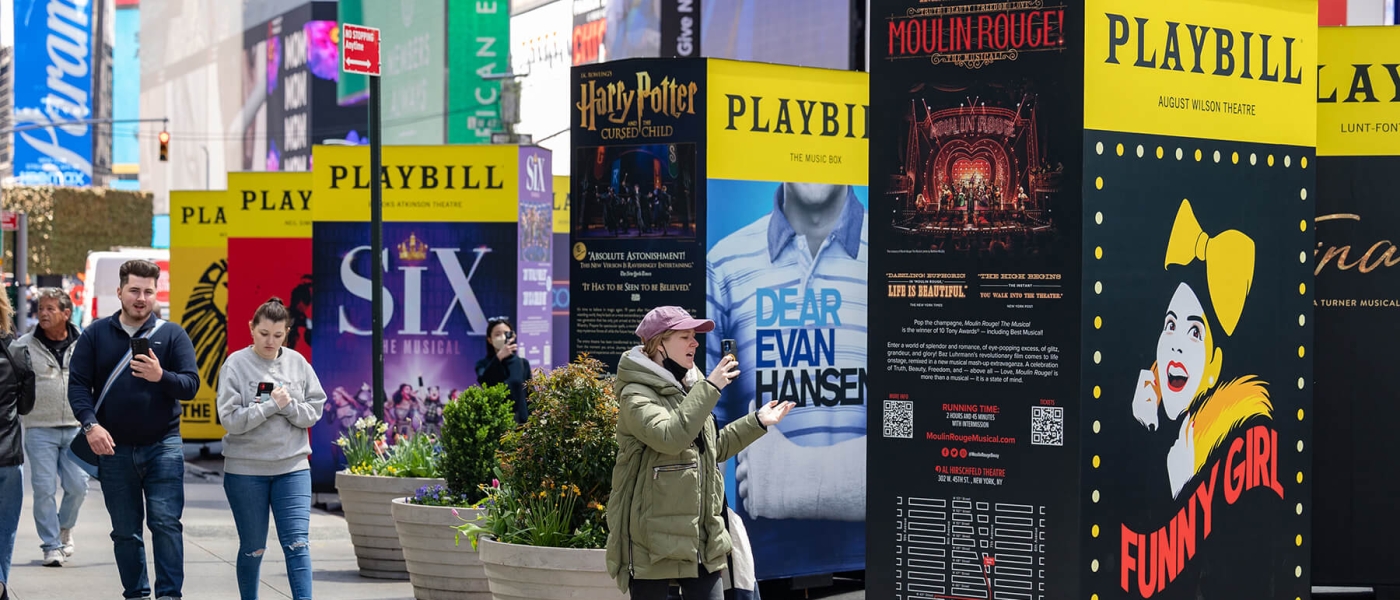 A visitor taking photos of the Broadway Grand Gallery, with giant Playbills for The Lion King, MJ, SIX, Harry Potter, Dear Evan Hansen, Moulin Rouge, Funny Girl, and Tina visible