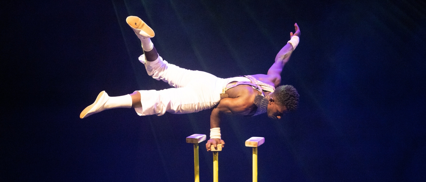 A Black man lying horizontal in the air, holding himself up with all his weight on a single hand balancing on a pole