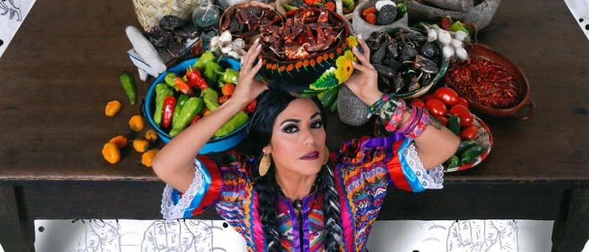 Lila Downs in a colorful outfit and braids, sitting in front of a table filled with bowls of peppers, reaching back with her hands