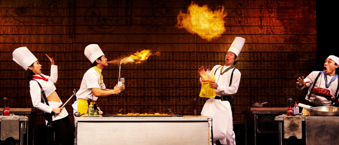 Four chefs onstage with a fry station between them. One chef is breathing fire as the other three chefs react in shock