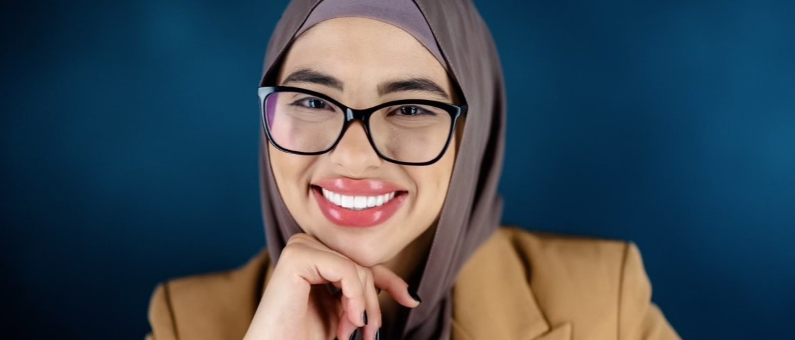 Nina Kharoufeh wearing glasses and a hijab, smiling at the camera with her chin on her hand