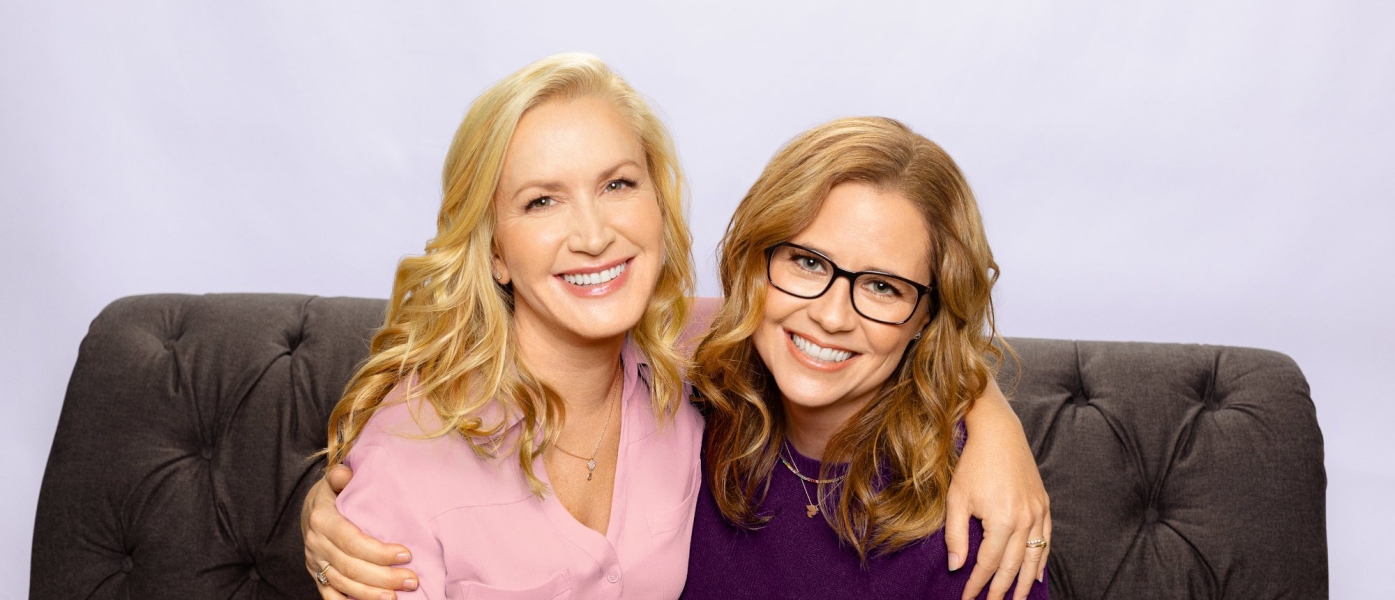 Angela Kinsey and Jenna Fischer sitting on a couch with their arms around each others shoulders
