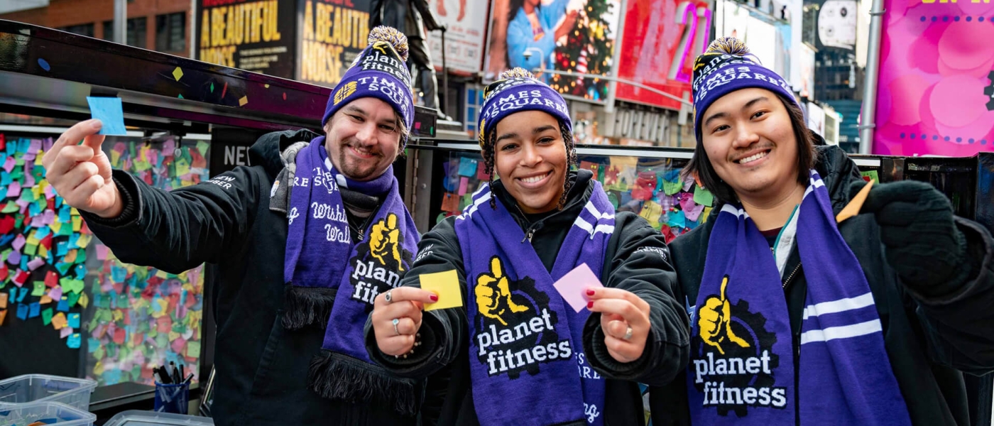 Three people in Planet Fitness hats and scarves, holding up pieces of confetti in front of the Wishing Wall.