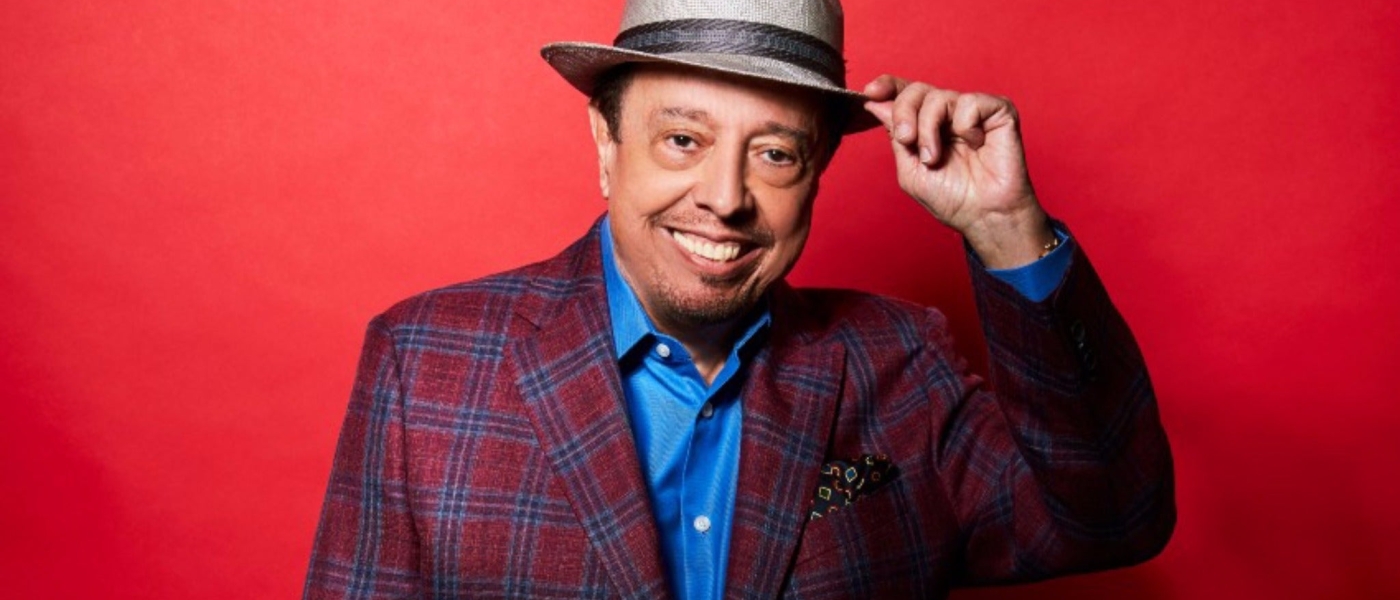 Sergio Mendes tipping his hat at the camera against a red background