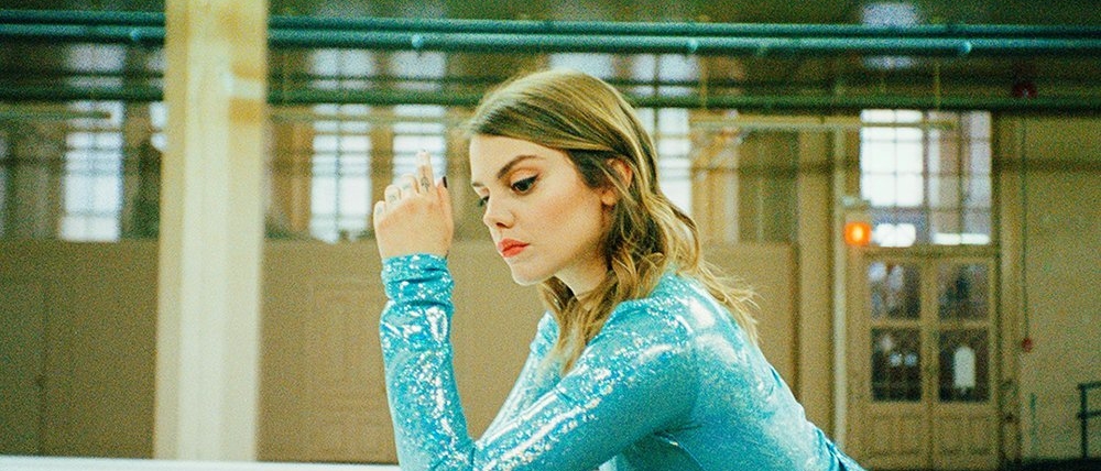 Coeur De Pirate wearing a shiny long-sleeved blue dress leaning on the edge of a white piano