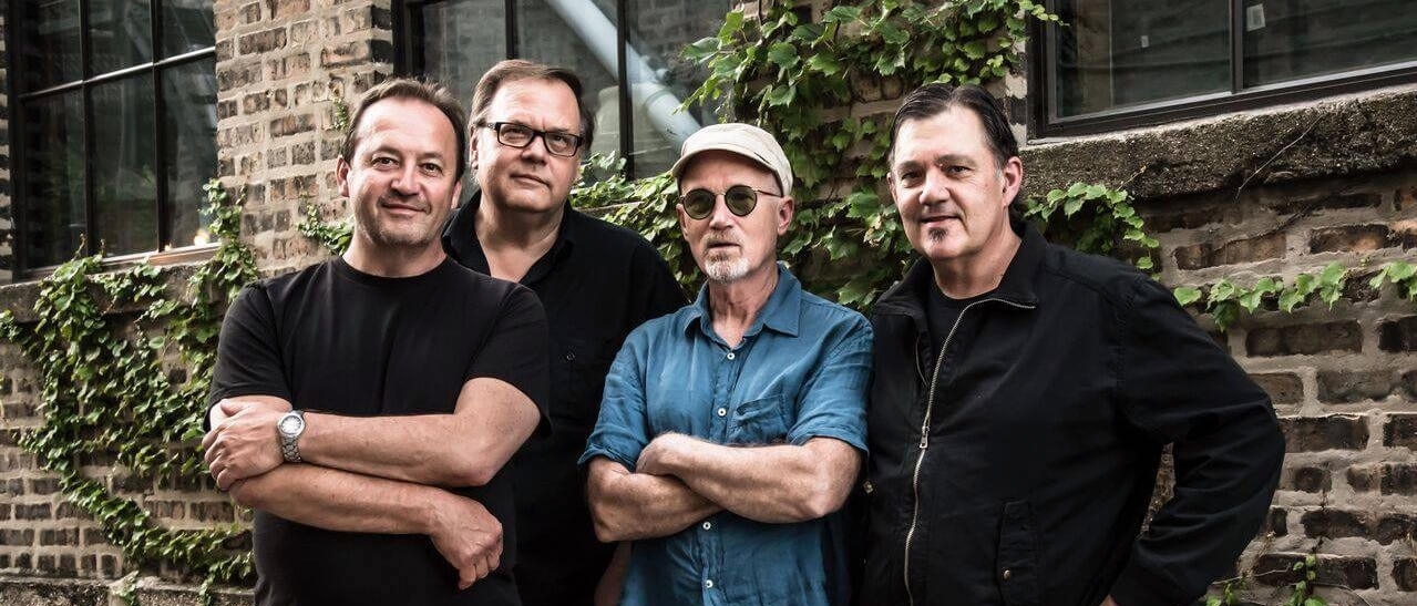 The Smithereens standing outside in front of an ivy-strewn brick building