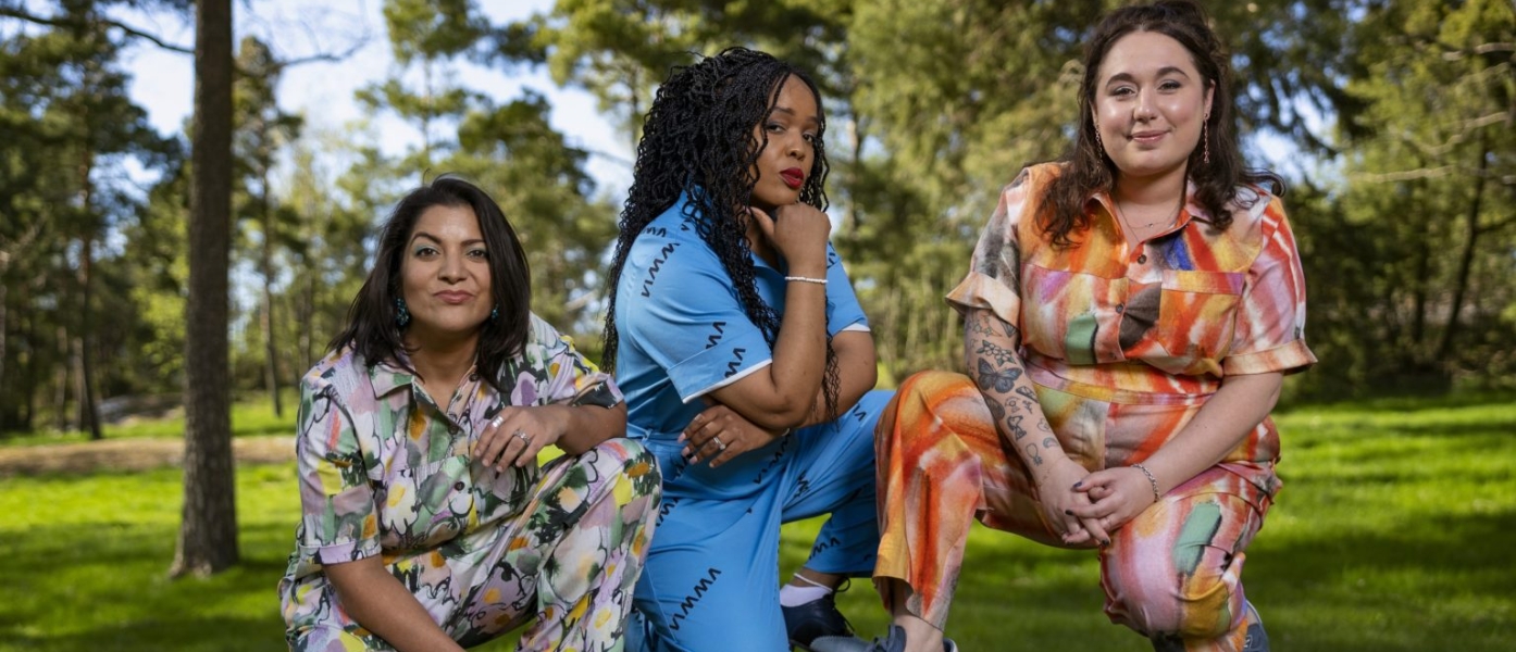 Aditi Mittal, Tumi Morake, and Liza Treyger wearing colorful patterned shirts and pants while crouching on a rock in a park