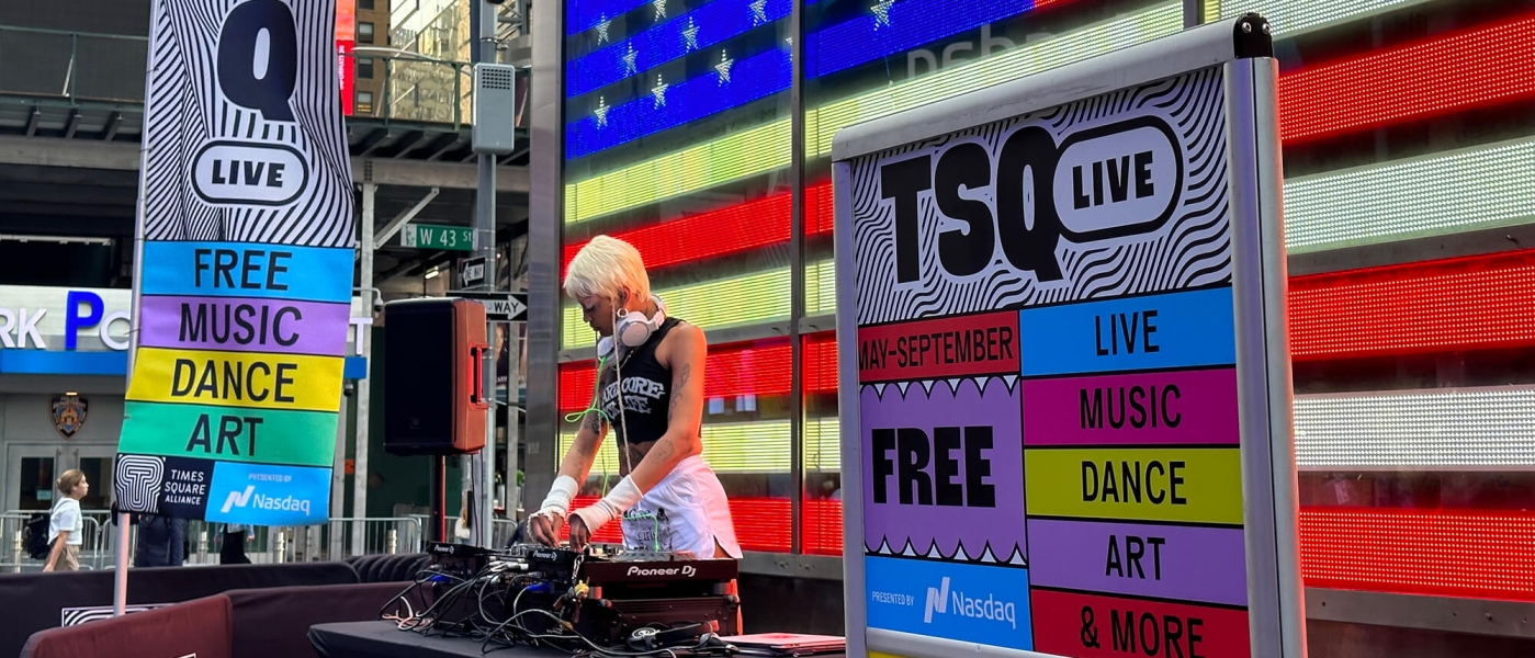 A DJ performing in Times Square in front of the giant LED American flag. Signs next to the DJ advertise TSQ LIVE.
