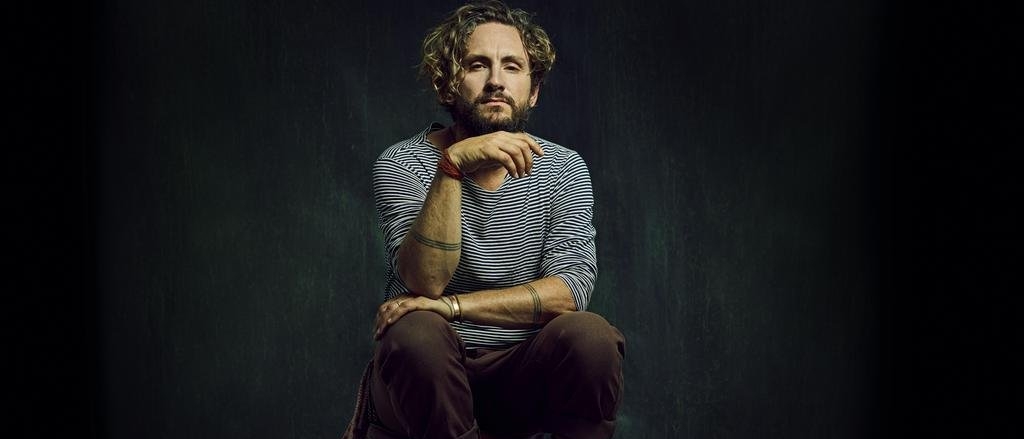 John Butler sitting on a stool with his elbow on his knee and his hand under his chin