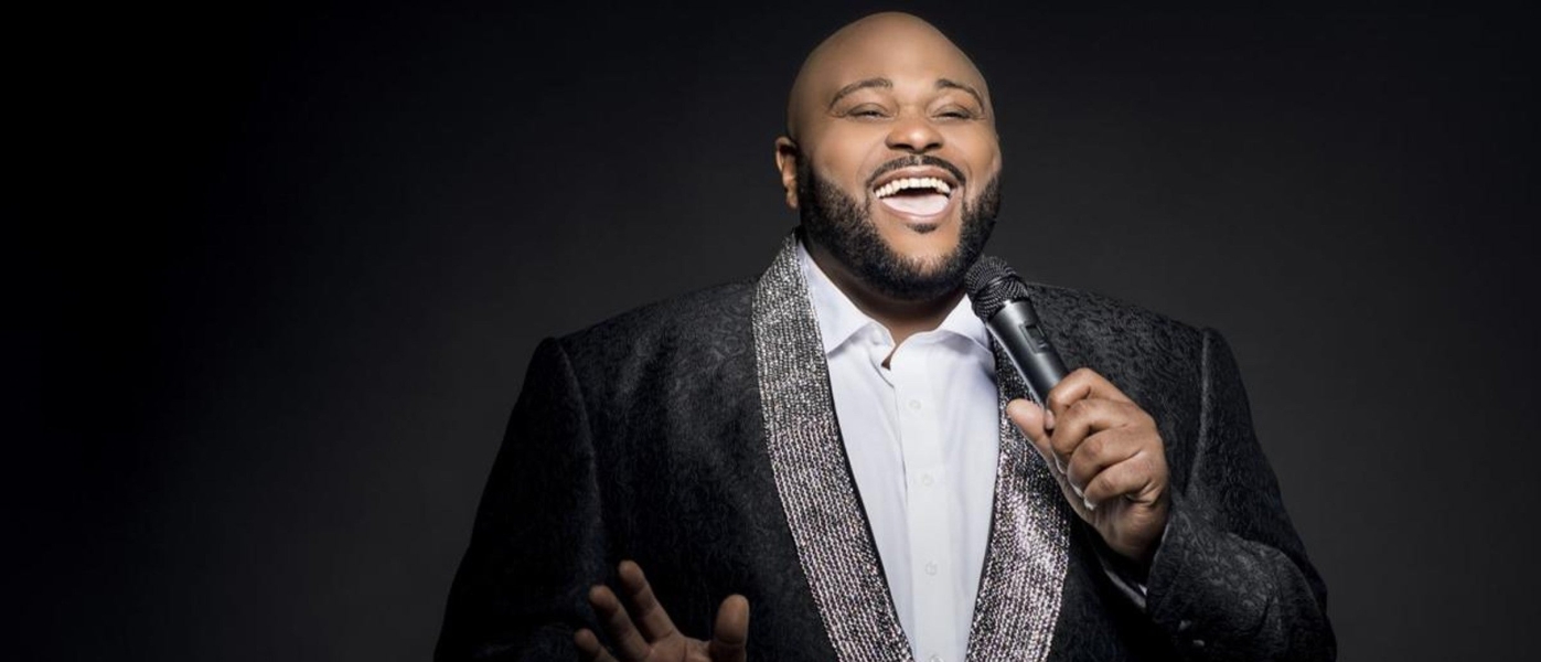 Ruben Studdard wearing a black suit with a silver shawl lapel. He is singing happily into a microphone.