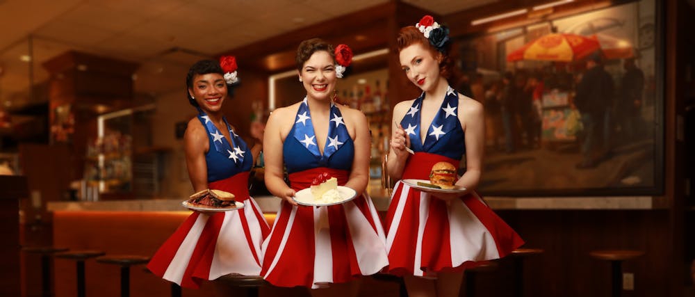 Three women in 1940s-style USO show troupe outfits in red, white and blue, each carrying a plate with a sandwich, a burger, or a slice of cheesecake