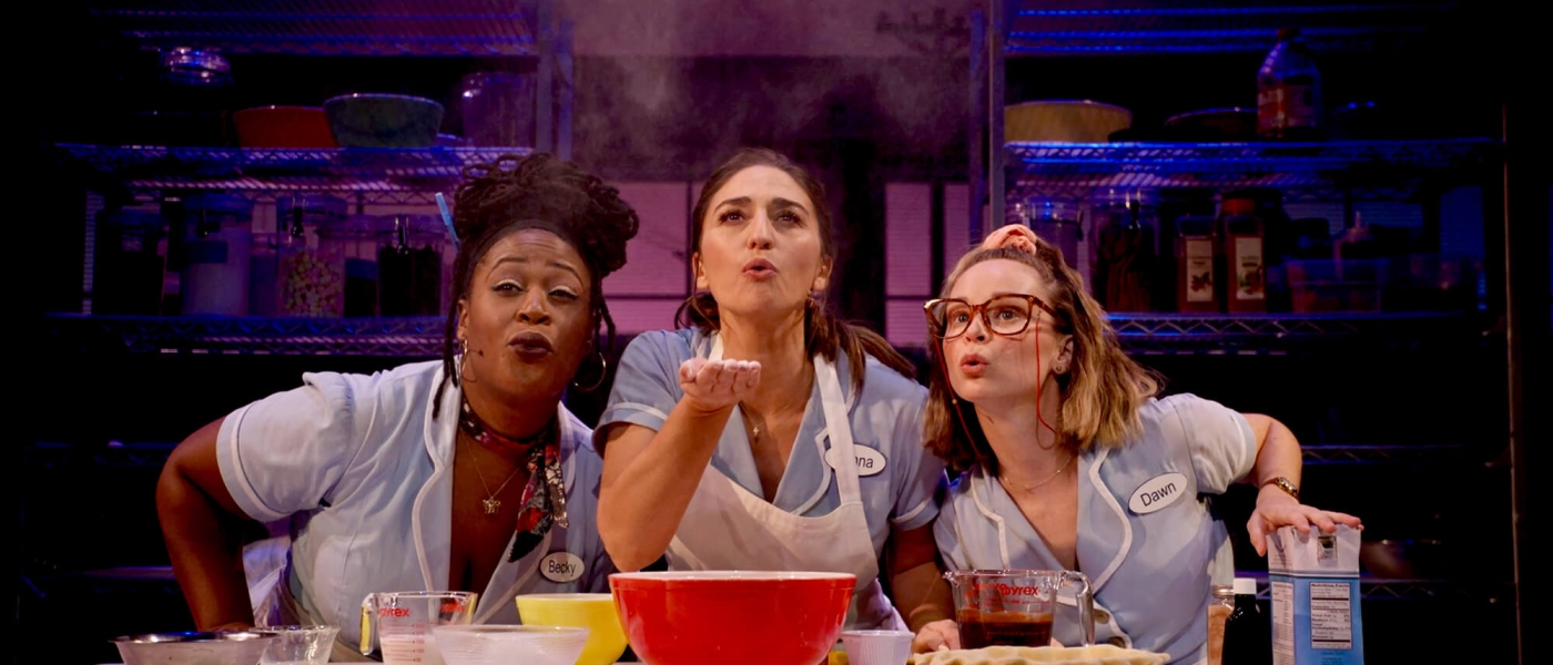 Charity Angél Dawson, Sara Bareilles and Caitlin Houlahan in 'Waitress, the Musical - Live on Broadway!', leaning towards each other over a table covered in baking supplies. Sara Bareilles, in the center, holds a handful of flour which she has just blown so it floats in the air