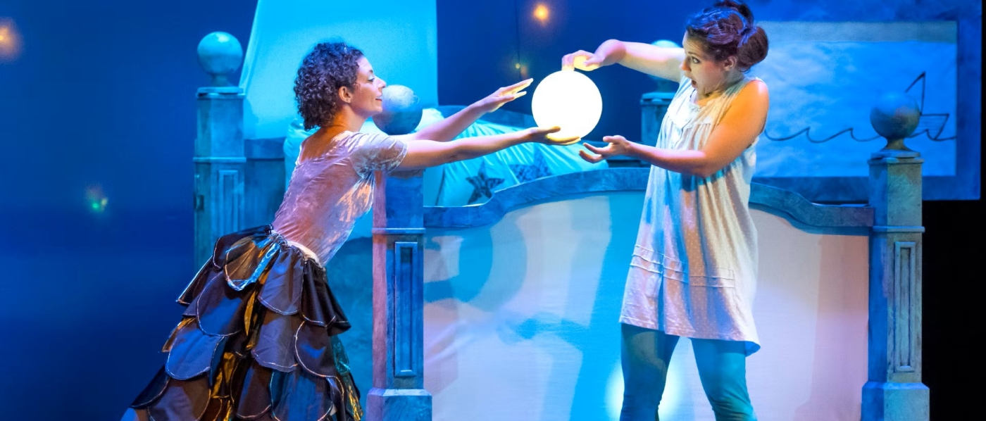 On a stage set as a bedroom, an actress dressed as a fairy and an actress dressed as a young girl hold a glowing orb between them. The young girl looks very surprised