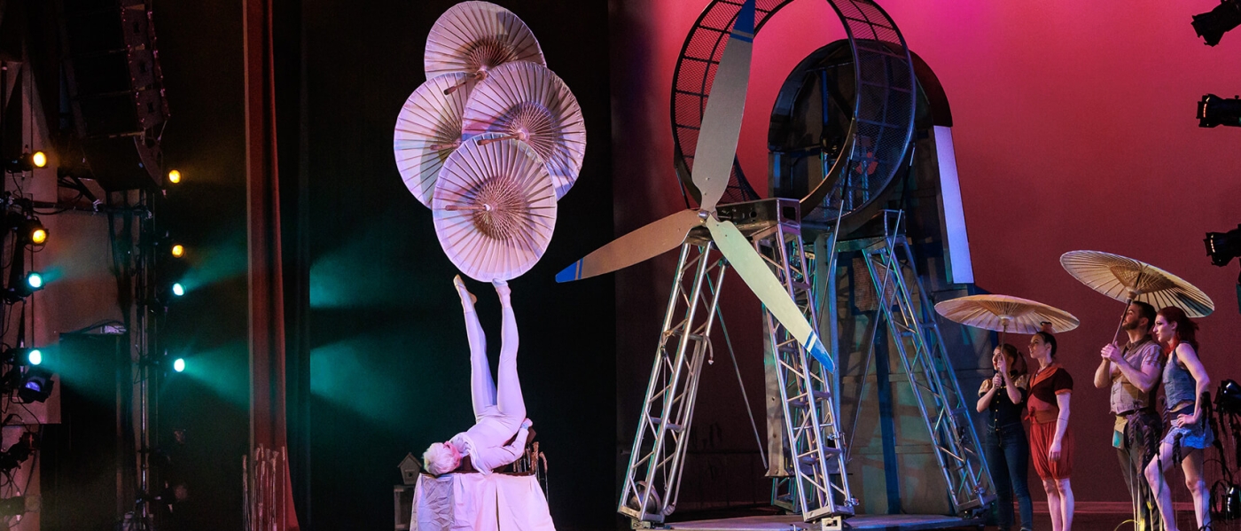 A performer wearing all white from Cirque Mechanics lies on their back with their legs up in the air, balancing four open white parasols on one foot