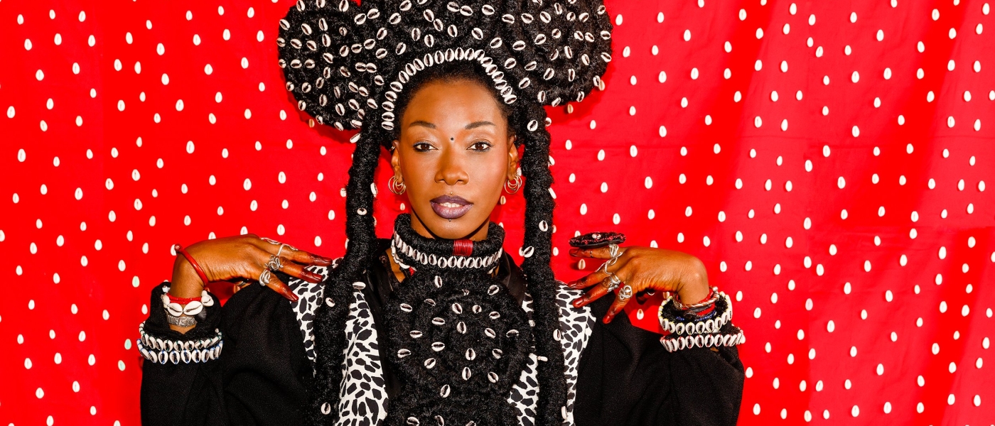 Fatoumata Diawara with an elaborate coiled hairstyle covered in puka shells in front of a white and red background