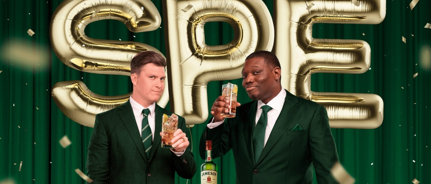 Colin Jost and Michael Che holding glasses with a bottle of Jameson and balloons reading "SPE" behind them