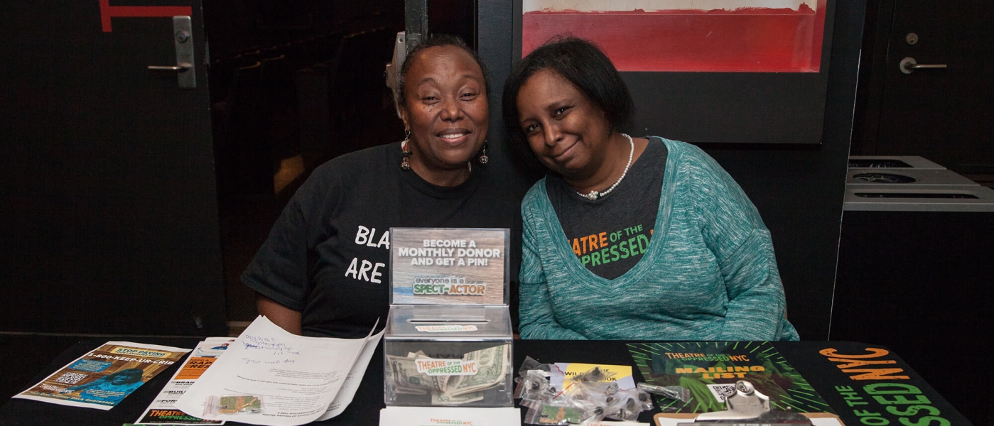 Two Theatre of the Oppressed NYC volunteers sitting at a table covered in informational material