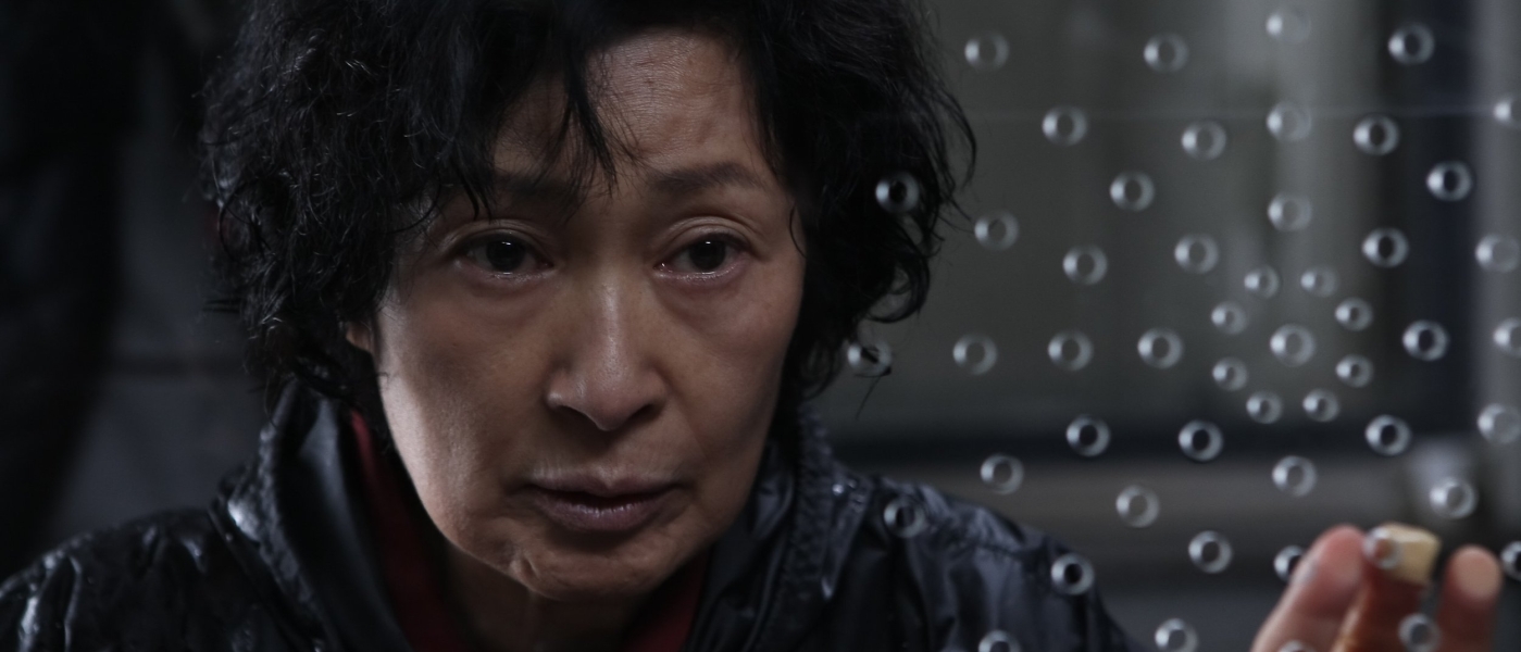 A still from Bong Joon-Ho’s movie "Mother," showing an older Asian woman looking through a sheet of plexiglass with small holes in it