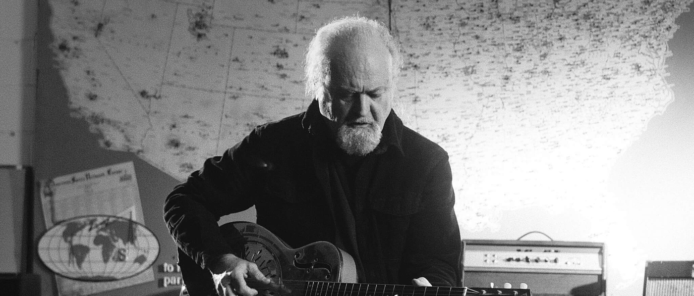 A black and white photo of Tinsley Ellis sitting on a stool playing a steel guitar