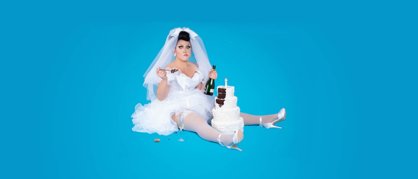 BenDeLaCreme sitting on the ground in a white bridal gown and veil, eating a slice of cake