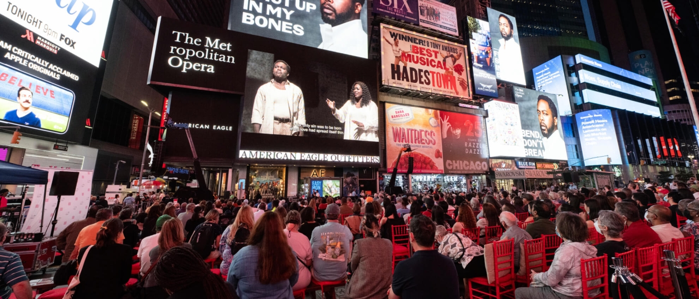 A photo from Met Opera Opening Night 2021 in Times Square, featuring a large number of people sitting on red chairs watching the opera Fire Shut Up in My Bones on the American Eagle screen