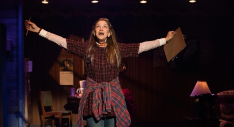 Victoria Clark in Kimberly Akimbo. She looks wrinkled but is dressed youthfully in jeans with a plaid flannel tied around her waist, and has her arms outspread