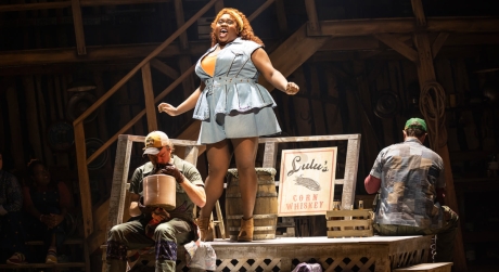 Alex Newell stands on a wooden platform on stage in Shucked on Broadway, next to a sign reading 