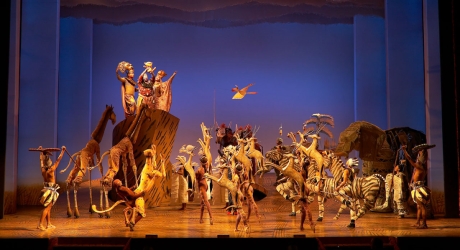 Wooden animal puppets and actors gather around Pride Rock as the actors playing Mufasa, Sarabi, and Rafiki lift the young Simba in this scene from The Lion King on Broadway