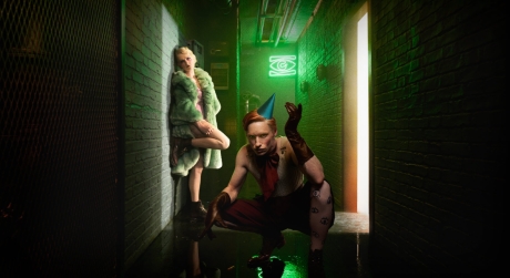 Eddie Redmayne and Gayle Rankin in a dimly-lit hallway. She wears a fur coat and boots and he wears a party hat, gloves, shorts, and a red bow around his neck